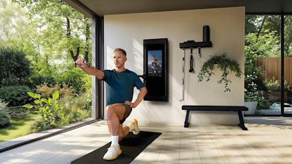 A man on a exercise mat down on one knee using the Tonal machine in a fitness room with floor to ceiling windows with views of surrounding gardens. 