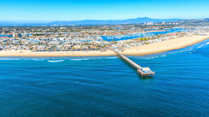 Aerial view of an Orange County beach with a long pier, marina, and urban coastline, perfect for showcasing the coastal setting of an autumn waterfront dinner. The image conveys a vibrant, seaside community atmosphere under a clear blue sky, ideal for hosting a memorable seafood meal by the water.