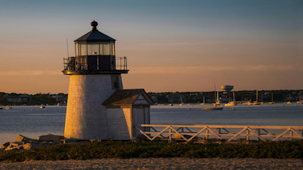A stunning sunset view of a Nantucket lighthouse on the coast, surrounded by calm waters and sailboats, perfectly capturing the serene and picturesque atmosphere of a spring or summer island escape.
