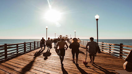 A panoramic view of a wooden pier extending into the ocean under a clear blue sky in San Diego, CA. People are walking and enjoying the sun, evoking the city's vibrant beachfront lifestyle and setting the mood for a culinary and wine festival by the coast.