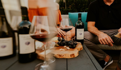 Close up of a table top that has a small charcuterie board and several bottles of Cakebread wine bottles with some glasses of red wine. 