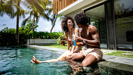 A couple sits on the edge of a backyard pool laughing with drinks in hand.