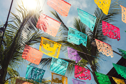 Colorful papel picado banners strung across the sky under bright sunlight with palm trees in the background in Punta de Mita, capturing the festive and vibrant spirit of Dia de los Muertos celebrations.