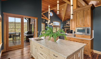 Alpine Ridge kitchen with large white island with modern light fixtures hanging above with glass double doors that lead out to an outdoor dining area with a large table and grill with views of the surrounding mountains. 