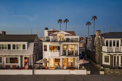 Sun-drenched Newport Beach beachfront home aglow at dusk, palm trees and rooftop terrace silhouetted against a deepening blue sky.