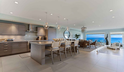 Kitchen area with large island and stainless steel appliances and dining area with circular table with floor to ceiling windows that look out to panoramic views of the coast.