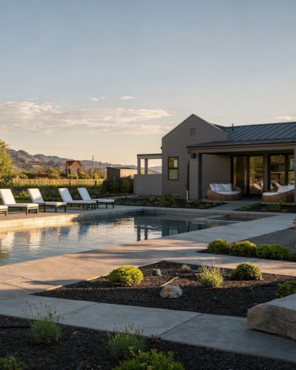 Modern dark gray estate with a stone-pillared patio, swimming pool, and lounge chairs under a clear blue sky.