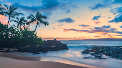 A serene Maui beach scene at sunset, with softly colored skies, silhouetted palms, gentle ocean waves and a tranquil shoreline, perfectly evoking the relaxing winter escape that luxury coastal homes near top winter festivals offer.
