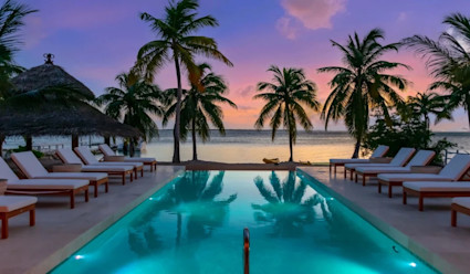 Pool area during a purple and pink sunset with rectangular pool illuminated with poolside lounge chairs and views of the ocean.