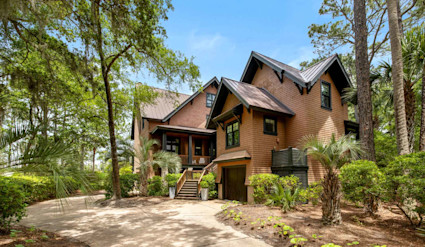Elegant cedar-shingle luxury home surrounded by palm trees and lush greenery, featuring a covered front porch and elevated entry in a private, wooded setting on Kiawah Island.