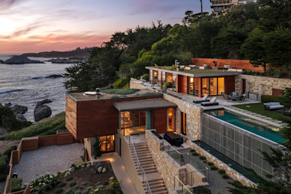 Modern cliffside home with infinity pool, glowing interiors, and rugged Pacific coastline at sunset.