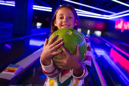 A stock image shows a smiling child holding a bowling ball in a bowling alley lit with neon lights.