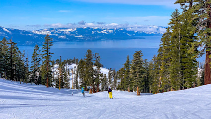 Skiers going down a slope toward the lake, surrounded by trees and blue sky.