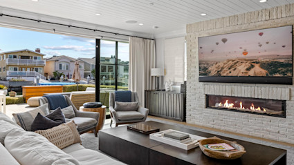 Living area with furniture arranged around a fireplace with a wall-mounted flat screen tv hanging above with sliding glass wall that leads out to furnished patio. 