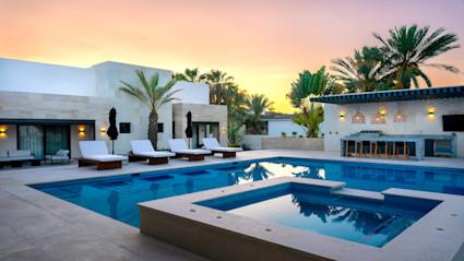 Solan pool area with hot tub with poolside lounge chairs and palm trees places around the area with a orange and yellow sunset in the sky. 