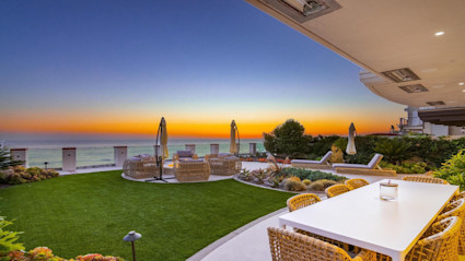A luxurious beachfront patio in San Diego, CA is set at sunset, featuring modern wicker furniture, umbrellas, and a large dining table overlooking the ocean. The well-manicured lawn and lush greenery, paired with the serene orange-and-blue sky, embody the essence of upscale outdoor entertaining in a luxury home.