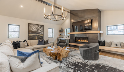 Living area with L-shaped couch, raw edge wooden coffee table, a wall-mounted flat screen tv hanging above a fireplace, and a chandelier hanging from the ceiling. 