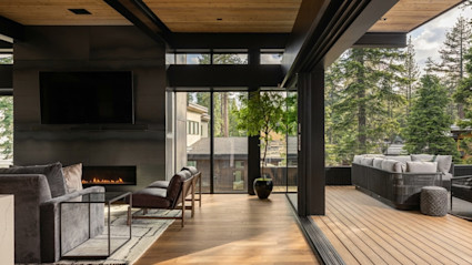 Modern living room with a lit fireplace opening through glass walls to a forest-view deck framed by towering pines.