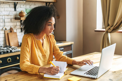 A woman in a yellow shirt sits in a kitchen with her laptop on the counter, likely enjoying a workcation.