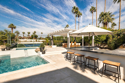 Resort-style backyard with a pool, swim-up bar and stools, shaded dining, and a tennis court framed by palms under blue skies.
