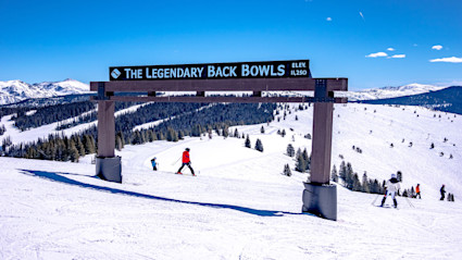 A pristine winter mountain scene in Vail, Colorado, with skiers traversing snowy slopes, surrounded by frosted trees and distant mountain peaks under a clear blue sky. This image captures the refined, serene ambiance that defines quiet luxury in winter vacation destinations.