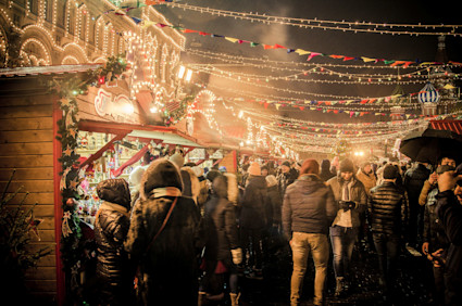 An outdoor holiday market with white lights illuminating the night sky as visitors crowd around stalls.