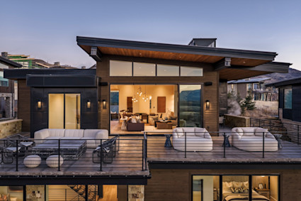 Modern mountain home at dusk, glowing interiors framed by glass walls and a furnished deck overlooking the foothills.