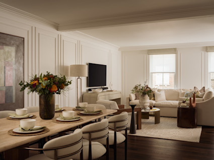 Elegant open dining and living area with a set table for six, fresh floral arrangements, paneled walls, and soft natural light.