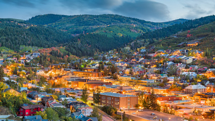An elevated twilight view of Park City, Utah, with glowing golden lights across a hillside town nestled in lush, pine-covered mountain ridges.