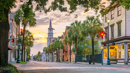 Historic street of Charleston lined by palm trees with the sun setting in a cloudy sky.