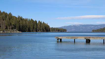 A tranquil wooden pier extends into the blue waters of Lake Tahoe, surrounded by pine forest and snow-capped peaks, evoking the serene promise of a spring break escape in a luxury lakefront setting.