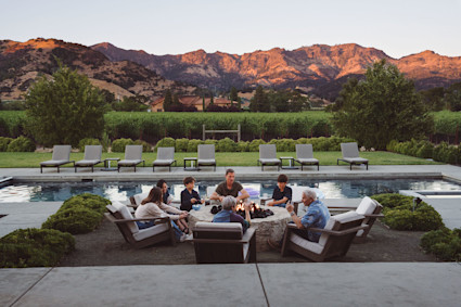 Family enjoying each other's company around a fire pit next to a pool with a mountain vista behind them. 