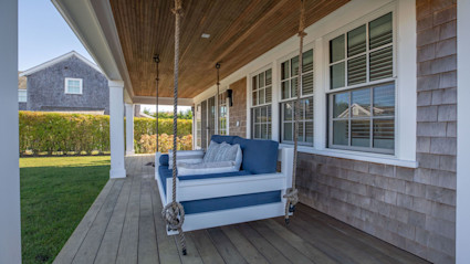 A cozy porch swing adorned with blue cushions and patterned pillows, set in a classic Nantucket shingle home, creates a welcoming space for families to gather, relax, and make memories together. The inviting, coastal ambiance reflects luxury and comfort—perfect for annual family traditions.