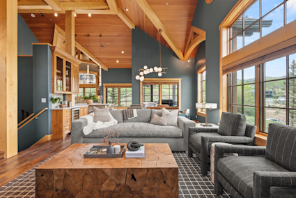 Sun-drenched mountain great room with vaulted timber ceilings, deep teal walls, plush gray seating, and sweeping views of the valley beyond.