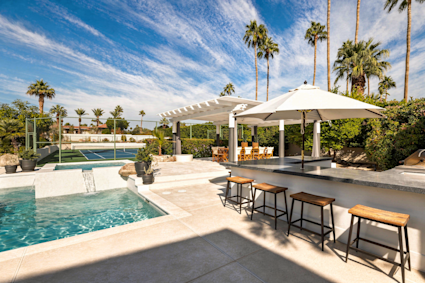 Desert backyard with a pool, shaded poolside bar, towering palms, and a tennis court under blue skies.