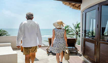 A couple walking with their backs to the camera across a patio towards the beach. 