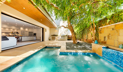A backyard pool shaded by a large tree with panoramic doors leading into the living room. 