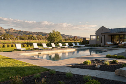 Wine country pool lined with white loungers, overlooking rolling vineyards and golden hills at sunset.