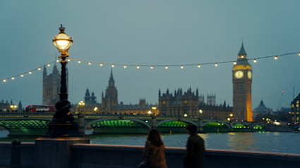 A twilight riverside view in London featuring the illuminated clock tower, festive string lights, and a red double-decker bus crossing a bridge. The scene captures the city's iconic architecture and vibrant atmosphere, ideal for those seeking the cultural richness of London's art and theatre season.