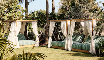 Outdoor cabanas with green striped cushions and white curtains surrounded by tropical palm trees on a sunny lawn.