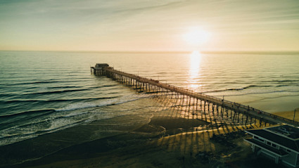A stunning sunset view of a pier stretching into the Pacific at Newport Beach, CA, with the golden hour lighting reflecting on the shoreline and the tranquil ocean. The long pier and open coastal expanse evoke the luxurious and relaxing essence of a Southern California coastal getaway.