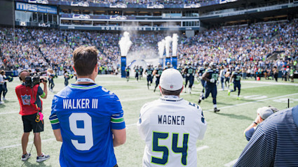 Two fans in football jerseys standing on the field at a stadium while the Seattle Seahawks football team rushes the field.
