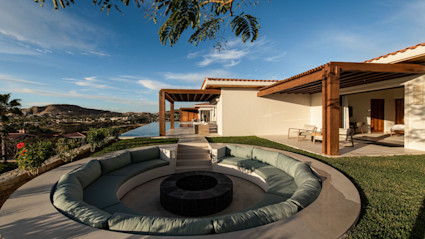 A luxurious villa in Cabo, Mexico featuring a sunken circular seating area with plush cushions around a fire pit, seamlessly blending indoor and outdoor living with an infinity pool and stunning views. This image captures the essence of relaxation and elegant design ideal for winter escapes.