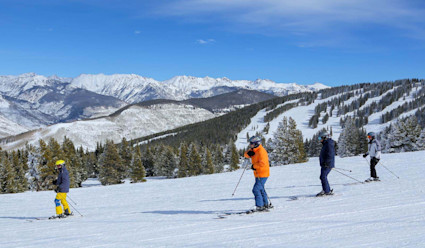 Four people skiing down a slope with snow covered mountains and a blue sky in the distance. 