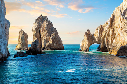 A breathtaking view of the iconic El Arco rock formation in Cabo San Lucas at sunrise or sunset, with dramatic cliffs rising from the blue ocean. The serene and picturesque scene captures the essence of a luxury winter beach retreat, invoking exclusivity and tropical beauty.