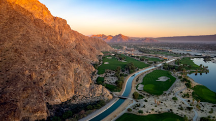 Aerial sunset view of a golf course in La Quinta, with dramatic mountains and water features, capturing the city’s reputation for picturesque and prestigious golf experiences.