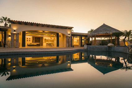 Hacienda-style villa glowing at dusk, its open living spaces and palapa reflected in a still infinity pool.