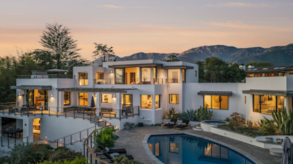 Modern multilevel white stucco estate at twilight with warm interior lighting, pool, and Santa Barbara mountain views.