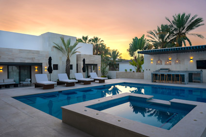 Pool and spa at dusk with chaise lounges, an outdoor bar, palm trees, and a Cabo sunset glowing behind a modern villa.