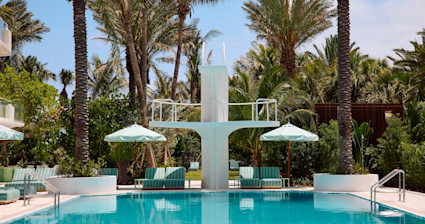 Resort pool with a white diving board, mint green lounge chairs, matching umbrellas, and lush palm trees on a sunny day.